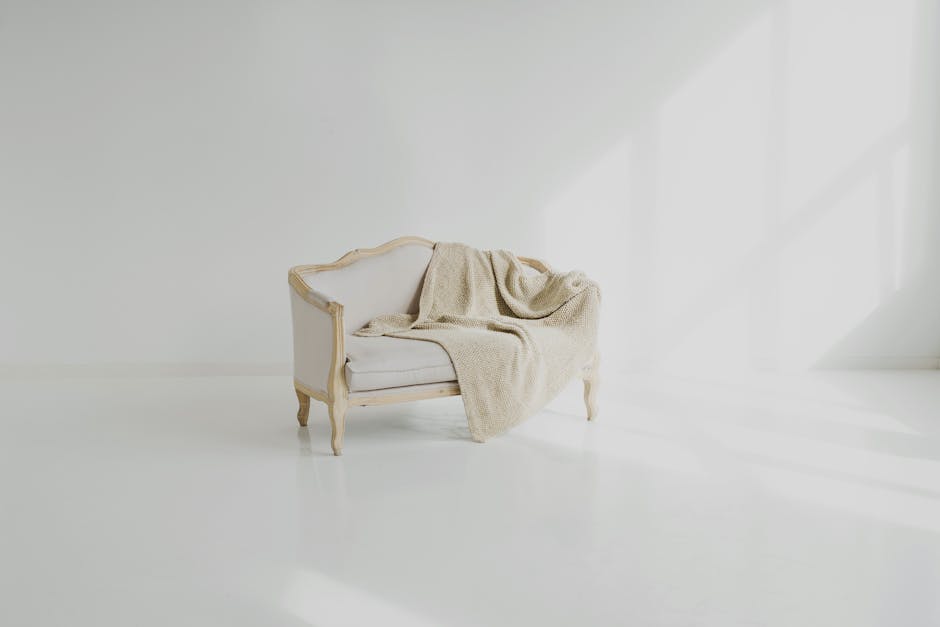 A white upholstered vintage armchair with wooden trim and curved legs, situated in a bright, minimalistic room with white walls and a clear, glossy floor. A beige, textured blanket is casually draped