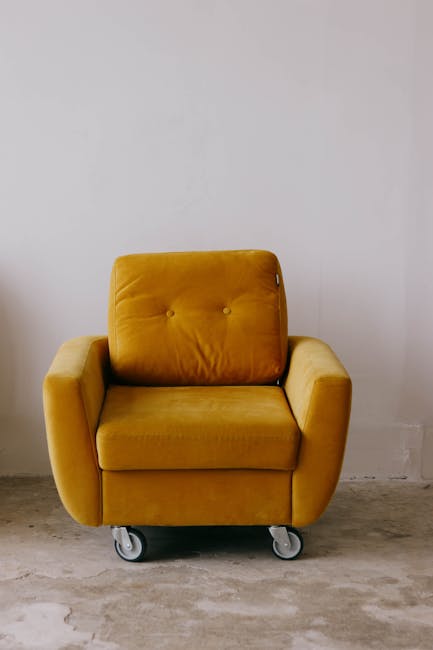 A white upholstered vintage armchair with wooden trim and curved legs, situated in a bright, minimalistic room with white walls and a clear, glossy floor. A beige, textured blanket is casually draped