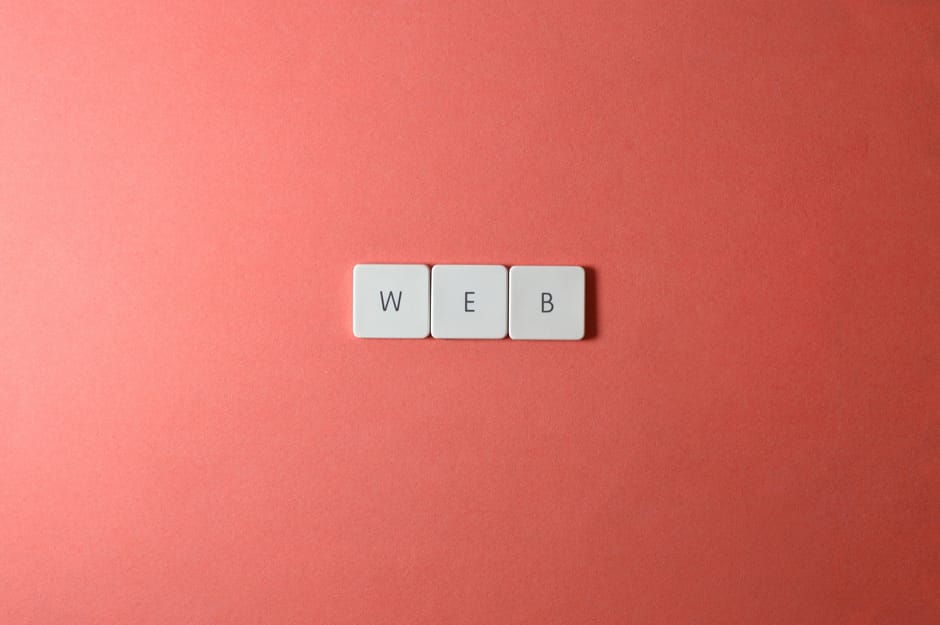 Close-up of three white square tiles with black letters spelling 'WEB' positioned evenly on a smooth, pastel pink surface, with the tiles aligned horizontally in the center of the image. The lighting